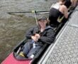 A young woman smiles while resting in a rowing shell on a river, with teammates preparing in the background.