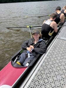 A young woman smiles while resting in a rowing shell on a river, with teammates preparing in the background.