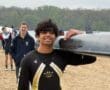 Smiling rower in a black and yellow uniform carries a boat on a sandy beach, with team members in the background.