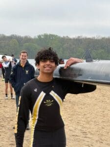 Smiling rower in a black and yellow uniform carries a boat on a sandy beach, with team members in the background.