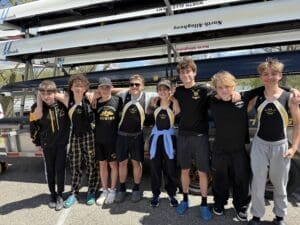 Eight rowing team members pose together in athletic gear, smiling with boats in the background on a sunny day.