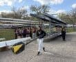High school rowers carry oars at a sports event, with boats and teammates in the background under a bright sky.