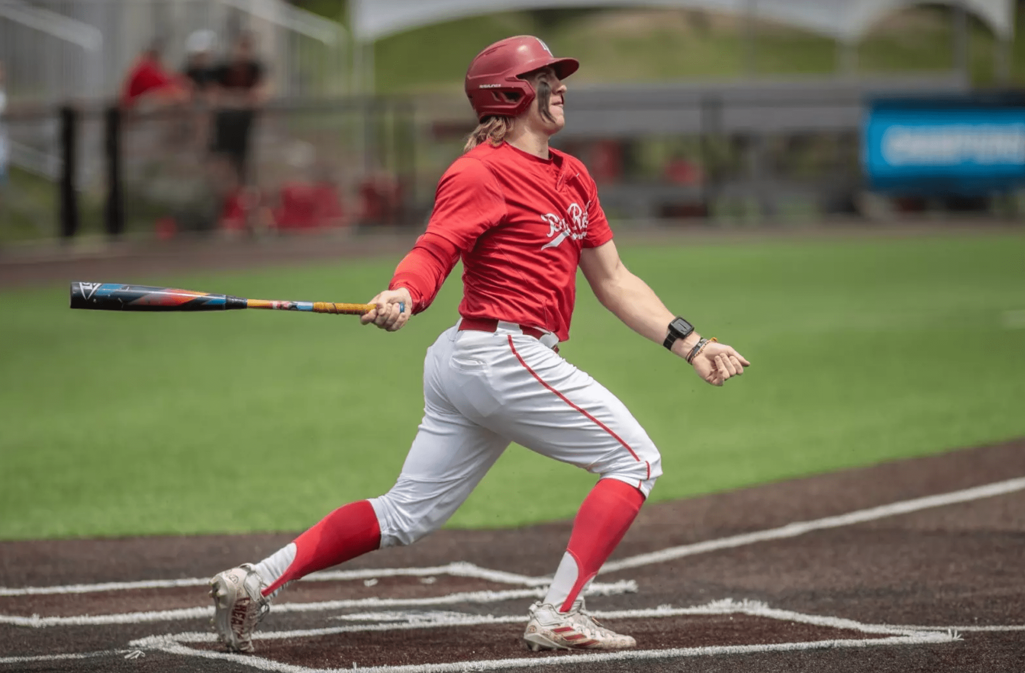 Baseball player swinging a bat on the field, wearing a red jersey and white pants. Action shot capturing athleticism.