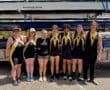 Rowing team poses with medals, celebrating their achievement at a competition near a trailer with rowing equipment.