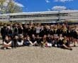 Rowing team celebrating awards outdoors, with gold medals, boats in the background, and a sunny sky above.