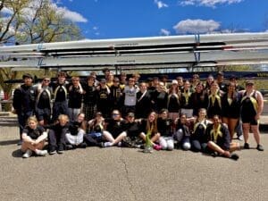 Rowing team celebrating awards outdoors, with gold medals, boats in the background, and a sunny sky above.
