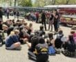 Rowing team gathers for instructions at an outdoor event under sunny skies, with boats and spectators in the background.