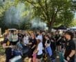 Participants gather in a wooded area, listening intently to a speaker amid smoke from nearby food vendors.