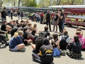 Rowing team gathers for instructions at an outdoor event under sunny skies, with boats and spectators in the background.