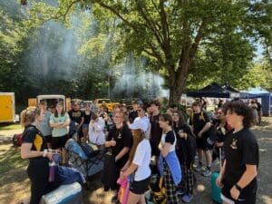 Participants gather in a wooded area, listening intently to a speaker amid smoke from nearby food vendors.