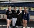 Four young women pose together by a trailer, showcasing teamwork and camaraderie in rowing attire.