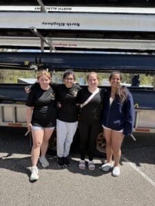 Four young women pose together by a trailer, showcasing teamwork and camaraderie in rowing attire.