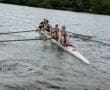 A rowing team in a sleek boat competes on a river under a partly cloudy sky, showcasing teamwork and athleticism.