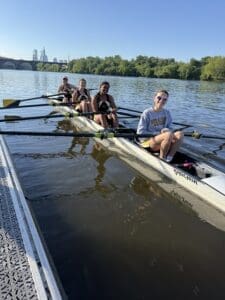Four female rowers in a boat on a river, enjoying a sunny day with trees and city skyline in the background.
