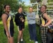 Four young female rowers in team gear enjoy a moment by their boat trailer, surrounded by greenery at a rowing event.