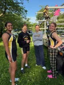 Four young female rowers in team gear enjoy a moment by their boat trailer, surrounded by greenery at a rowing event.
