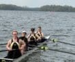 Rowing team of four women in athletic gear, smiling on a river with trees in the background on a cloudy day.