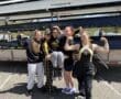 A group of young female rowers in team attire, celebrating by flexing muscles by their rowing boat trailer.