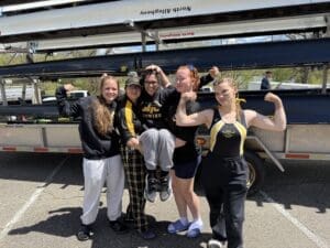 A group of young female rowers in team attire, celebrating by flexing muscles by their rowing boat trailer.