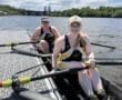 Two female rowers prepare on a dock, smiling with oars in hand, showcasing teamwork on a sunny day by the water.