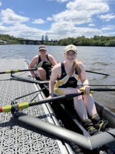 Two female rowers prepare on a dock, smiling with oars in hand, showcasing teamwork on a sunny day by the water.