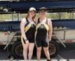 Two smiling young women in rowing attire stand by a boat trailer, ready for a day of rowing.