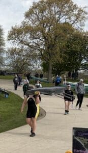 Rowers carrying boats along a pathway during a rowing event, with spectators and greenery in the background.