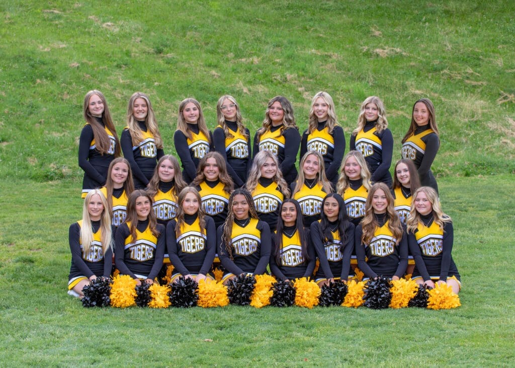 Cheer team photo with members in black and gold uniforms, holding pom-poms, posed on a grassy field.