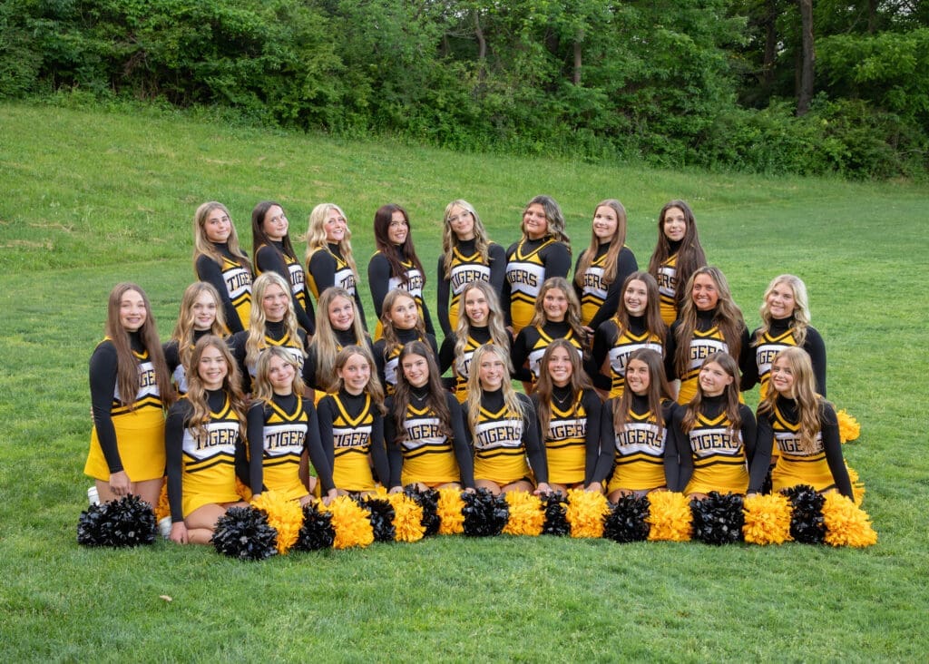 Cheerleaders in black and gold uniforms pose together outdoors, showcasing team spirit with pom-poms on the grass.