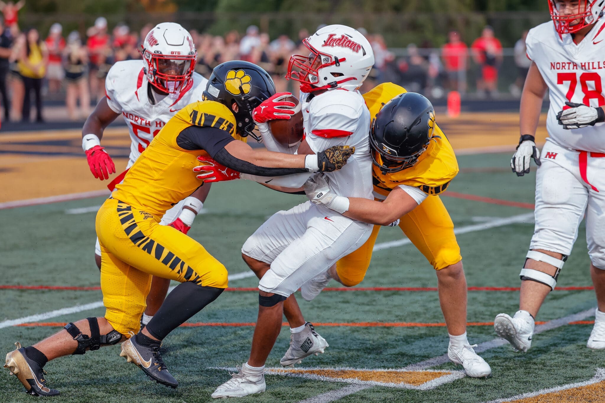 High school football action with players tackling an opponent while spectators cheer in the background.