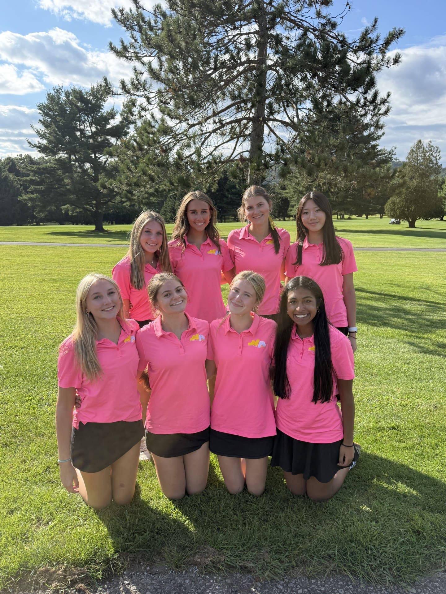 Group of eight girls in bright pink golf shirts posing outdoors on a sunny day, surrounded by green grass and trees.