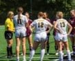 Girls' soccer teams huddle before the game, discussing strategy with officials on the field in sunny weather.