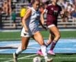 A female soccer player in white dribbles the ball, with another player in maroon close behind on a sunny field.