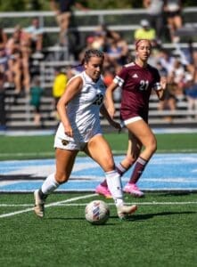 A female soccer player in white dribbles the ball, with another player in maroon close behind on a sunny field.