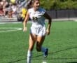 A female soccer player in a white jersey dribbles a ball on an artificial turf field during a sunny match.