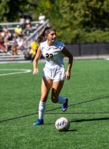 A female soccer player in a white jersey dribbles a ball on an artificial turf field during a sunny match.