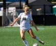 High school female soccer player in action, kicking the ball on a sunny field during a state championship match.