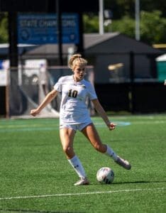 High school female soccer player in action, kicking the ball on a sunny field during a state championship match.