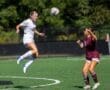 Two female soccer players on the field: one jumps to head the ball while the other prepares to defend.