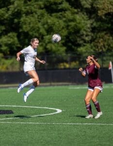 Two female soccer players on the field: one jumps to head the ball while the other prepares to defend.