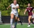 Soccer player in white jersey dribbles the ball on a sunny field, with teammates and opponents in motion behind her.