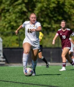 Soccer player in white jersey dribbles the ball on a sunny field, with teammates and opponents in motion behind her.