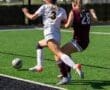 Two female soccer players compete for the ball on a green turf field during a match in bright sunlight.