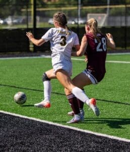 Two female soccer players compete for the ball on a green turf field during a match in bright sunlight.