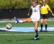 A female soccer player in action, kicking a ball on a sunny field with a referee in the background.