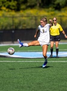 A female soccer player in action, kicking a ball on a sunny field with a referee in the background.