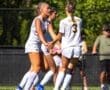 Three female soccer players in white uniforms celebrate on the field during a sunny game, showcasing teamwork and energy.