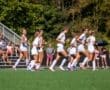 A group of female soccer players running together on a field, with spectators cheering in the background.