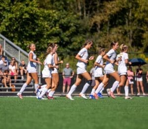 A group of female soccer players running together on a field, with spectators cheering in the background.