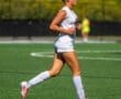Young female soccer player running on a field, wearing a white jersey and pink cleats, focused on her game.